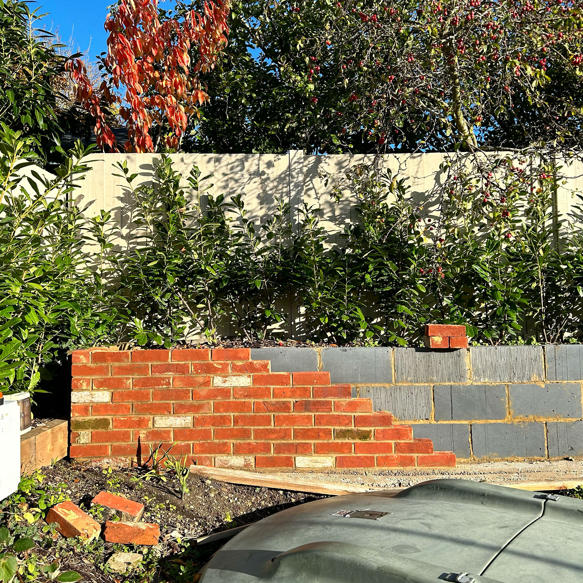 Garden with breezeblock wall being covered in red bricks, in front of fence and hedge area
