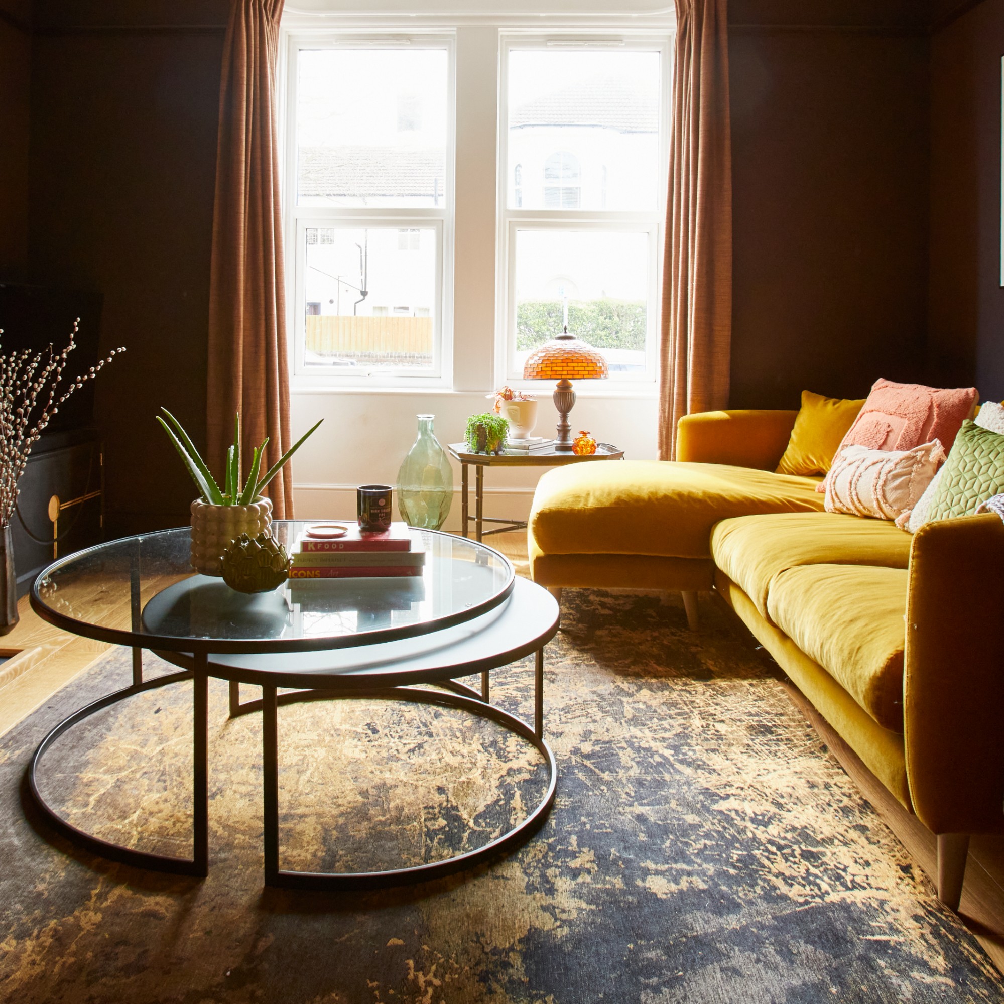 A dark living room with a yellow chaise sofa, a round nesting coffee table and a vintage amber glass table lamp