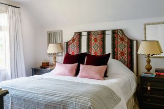 bedroom with bright patterned paneled fabric bedhead and double bed with pink cushions. striped bedspread and vintage bedside chests with table lamps and scalloped lampshades