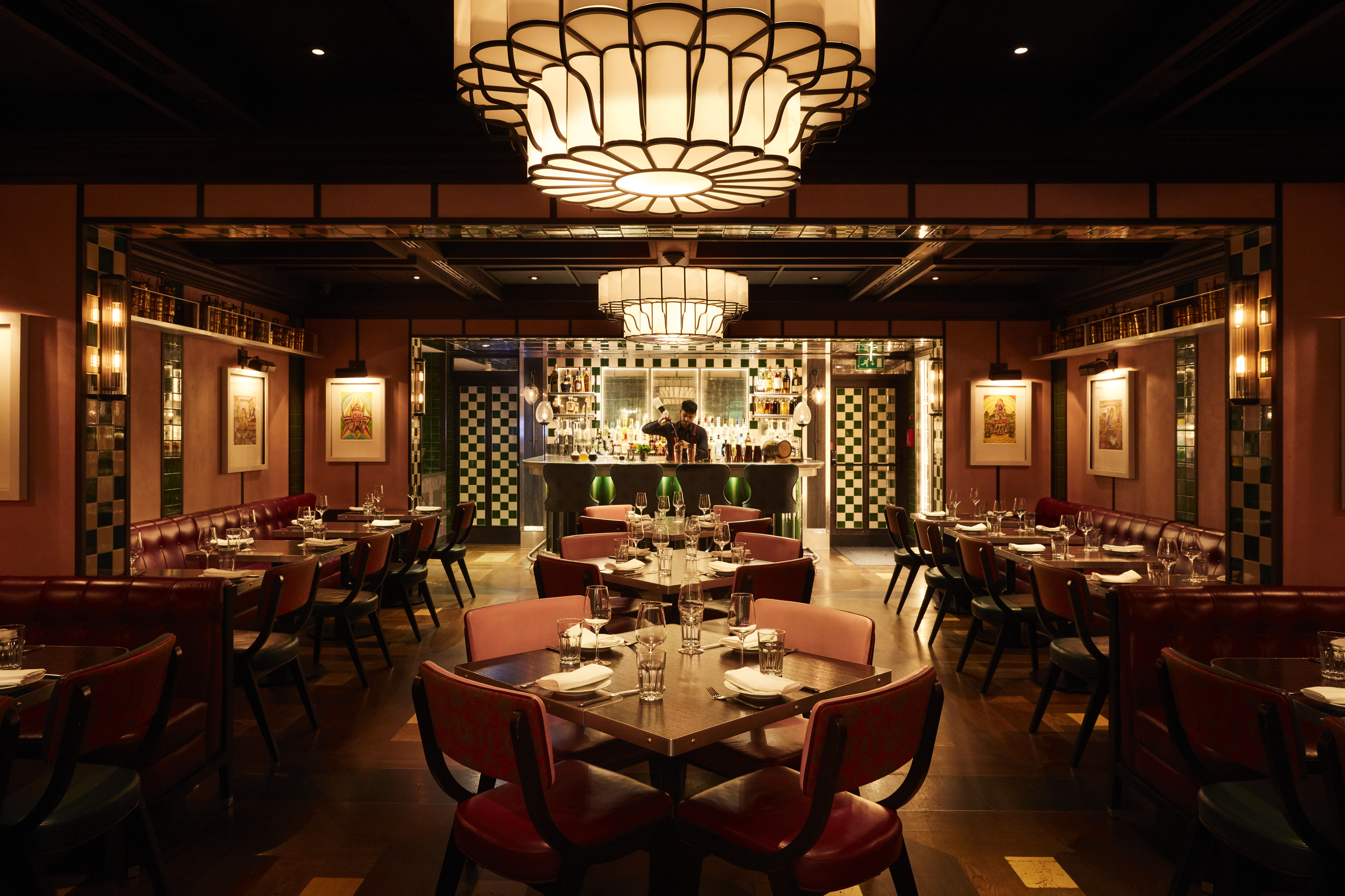 A vibrantly decorated Indian restaurant with green and while tilework, argilla-tinted walls, leathery seats, a large, pending chandelier hanging above a central, lavish bar, and a railway carriage-inspired atmosphere.