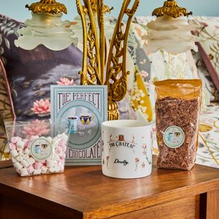 a wooden bedside table with a gold ornamental lamp with frosted petal shades on top, alongside mini marshmallows, chocoloate pieces, a personalised mug and a sign saying the perfect hot chocolate