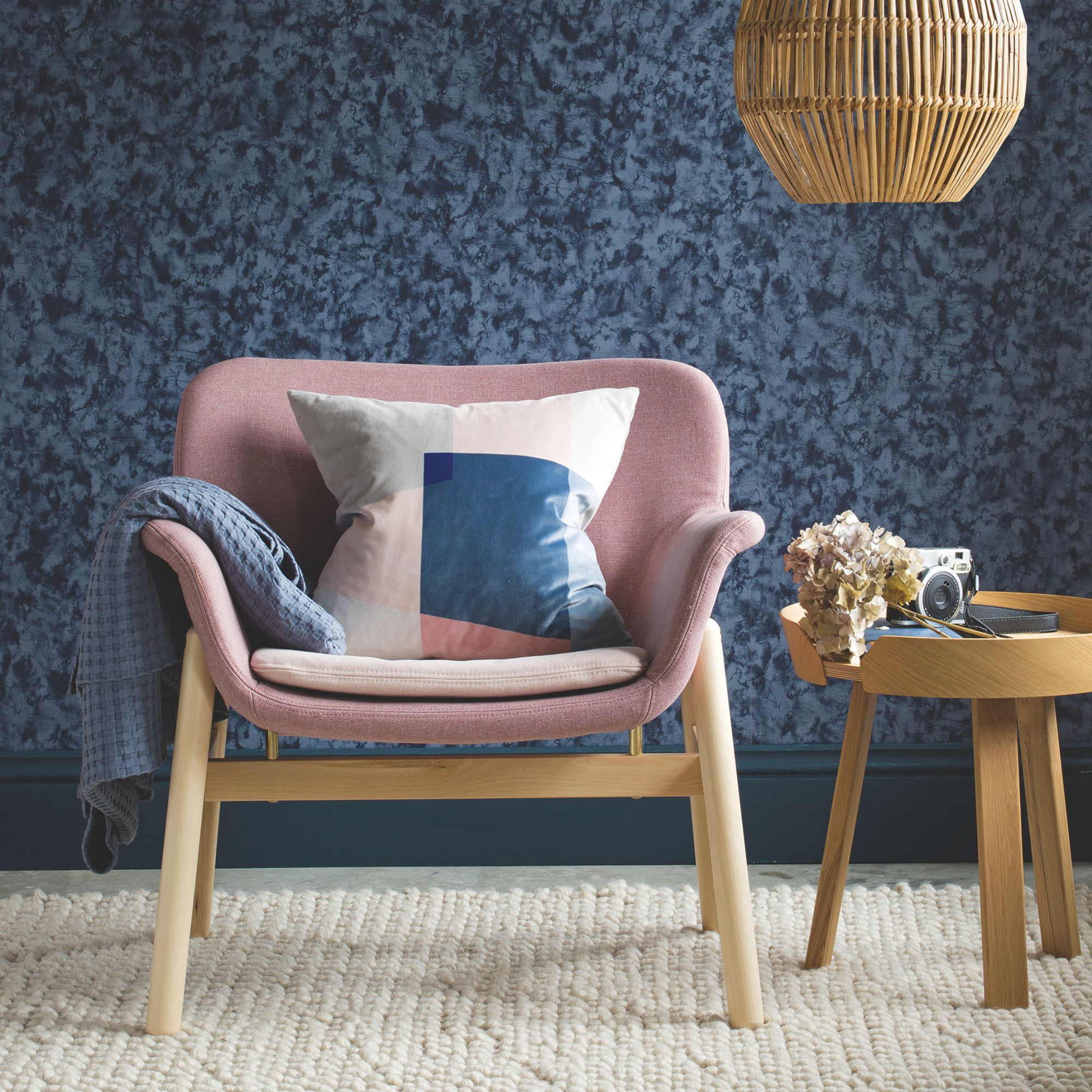 A dark blue wallpaper wall behind a blush pink chair with blue accent cushion