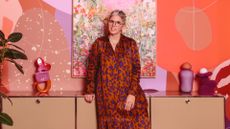 A woman in a red and purple maxi dress and dark glasses stands in front of a fancifully decorated wall in tones of orange, earth, pink, and purple, right before a floral canvas. She sits on a cabinet decorated with softly shaped objects in the same shades.