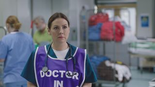 Stevie Nash looks stunned in the ED. She's wearing her blue scrubs with a purple bib with the words 'DOCTOR IN CHARGE'.