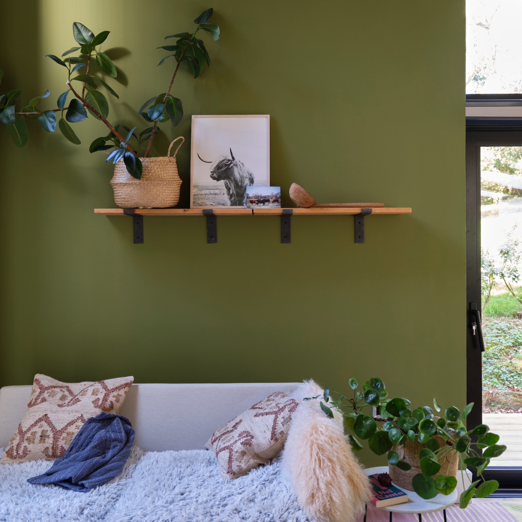A living room painted in Farrow &amp;amp; Ball's Sap Green shade with a cream sofa with textured soft furnishings and a shelf above
