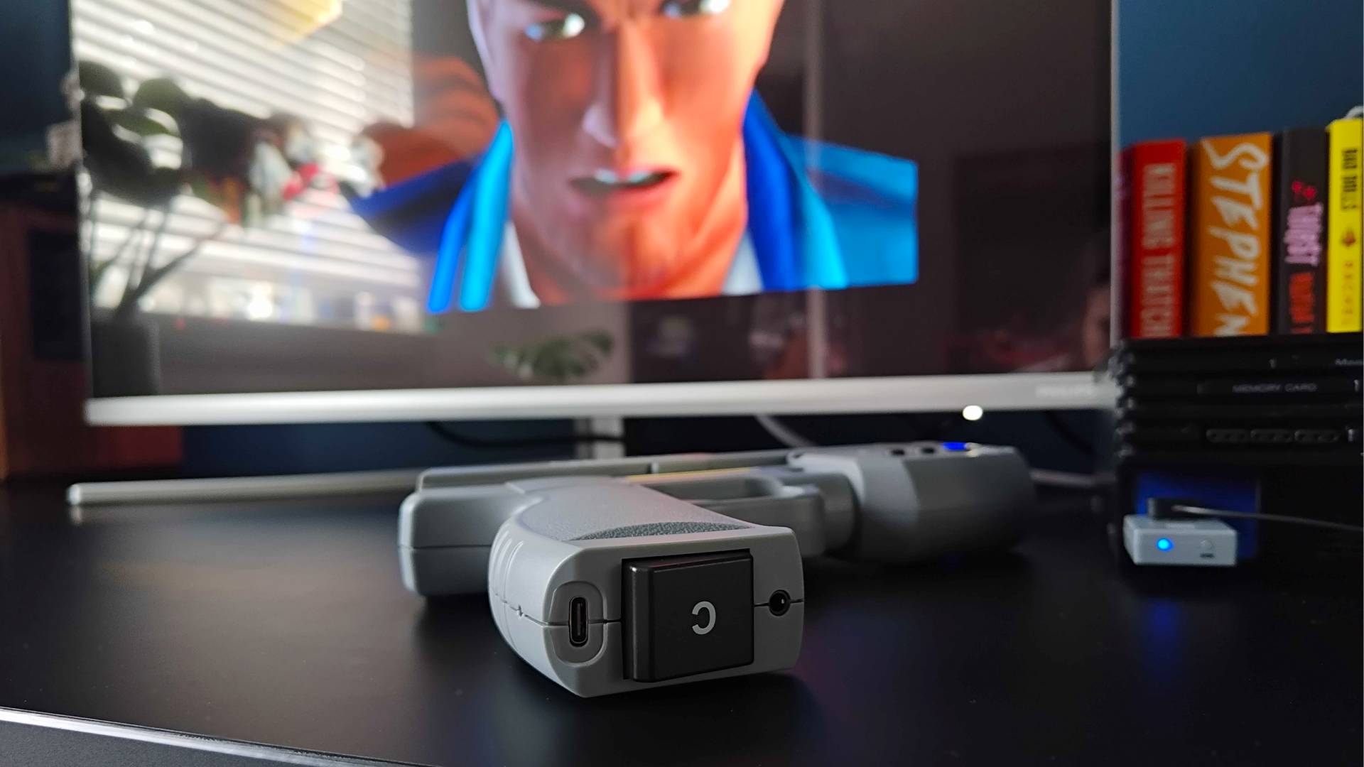 Intec Time Breaker Lightgun lying on black desk with bottom C button facing camera next to PS2 and monitor with Time Crisis 2 intro in backdrop.