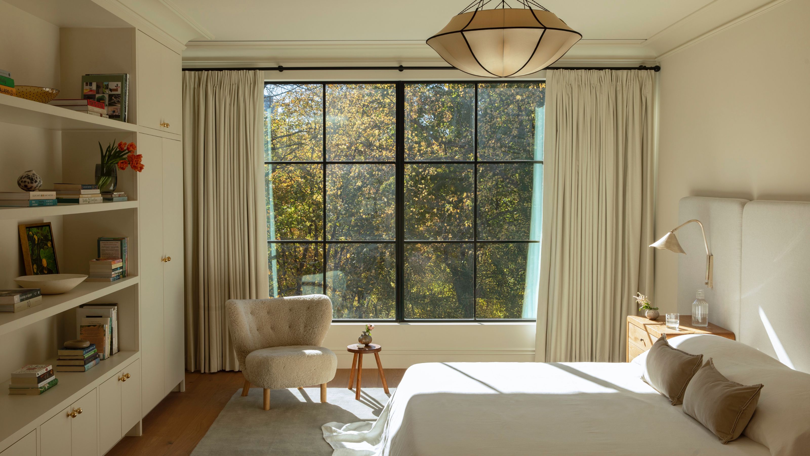 A bright and peaceful primary bedroom featuring a large black-framed window that looks out onto vibrant autumn trees. The room includes a bed with a tall, light-gray upholstered headboard and neutral bedding, a cozy cream boucl&amp;eacute; armchair by the window, and a floor-to-ceiling built-in shelving unit filled with books. A large, saucer-shaped pendant light hangs from the ceiling, casting a soft glow over the warm wood floors