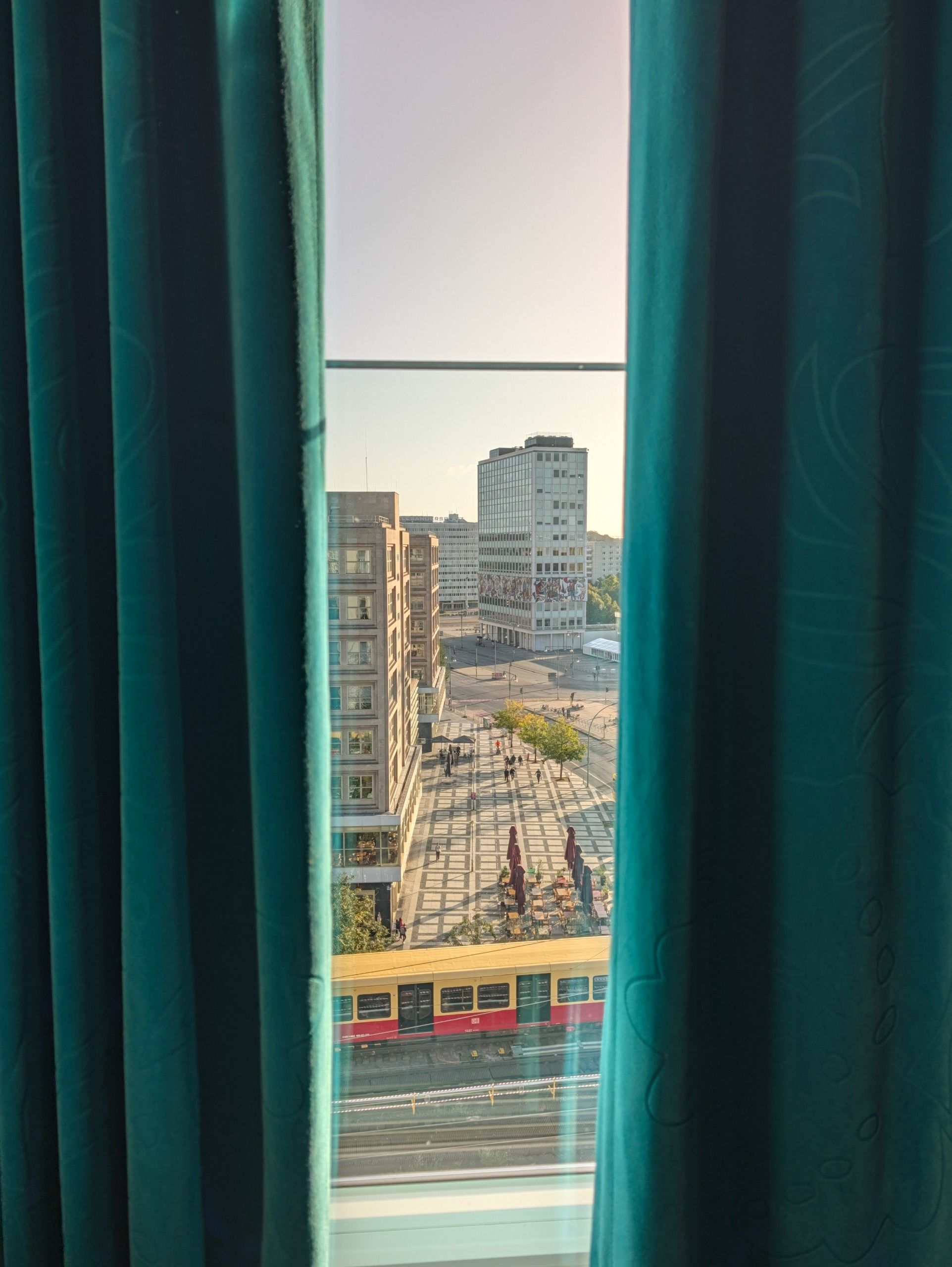Berlin city view framed by partially closed teal hotel curtains, with a train passing below.