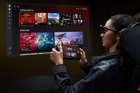 Woman wearing ROG XREAL AR glasses while holding a ROG Ally X20 handheld, projecting a large virtual gaming display featuring Xbox games and the Xbox interface.
