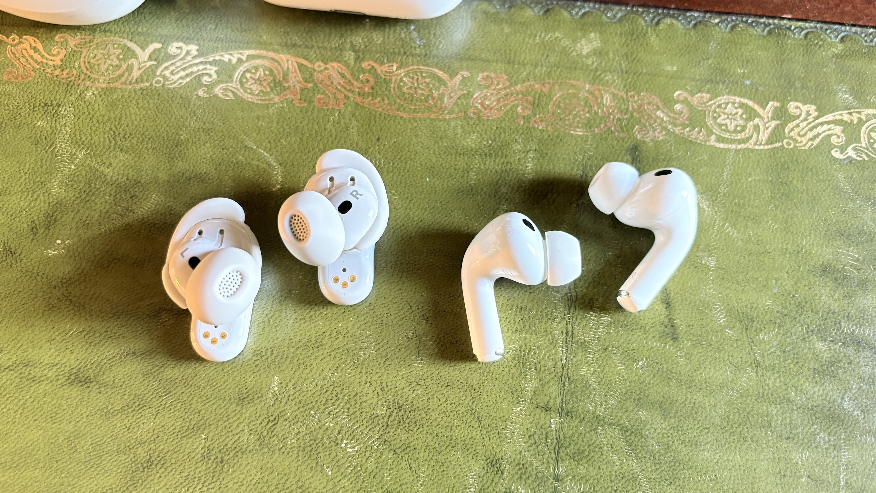A pair of Bose QuietComfort Ultra Earbuds (2nd Gen) next to a pair of AirPods Pro 3 on a green surface.