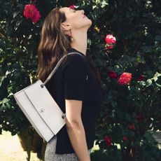 A woman stood side on to the camera with her head up and eyes closed in front of a rose bush holding a Tory Burch handbag on her shoulder