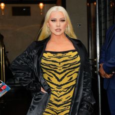 a white woman with platinum blonde hair being photographed in front of a building wearing a yellow animal print dress and a black silk jacket