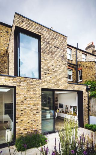 a significant brick building extension, with a large white kitchen inside and a small patio with plant on it outside
