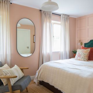 Pink bedroom with panelling behind the bed, a mirror on the wall, white curtains, and a blue accent chair