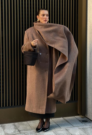 a photo of a woman standing on the sidewalk wearing gold earrings with a long tan coat with an oversized matching scarf styled with a suede bucket bag and black pointed kitten heels