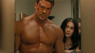 Alan Ritchson shirtless in Reacher while Agnez Mo stands behind him. 