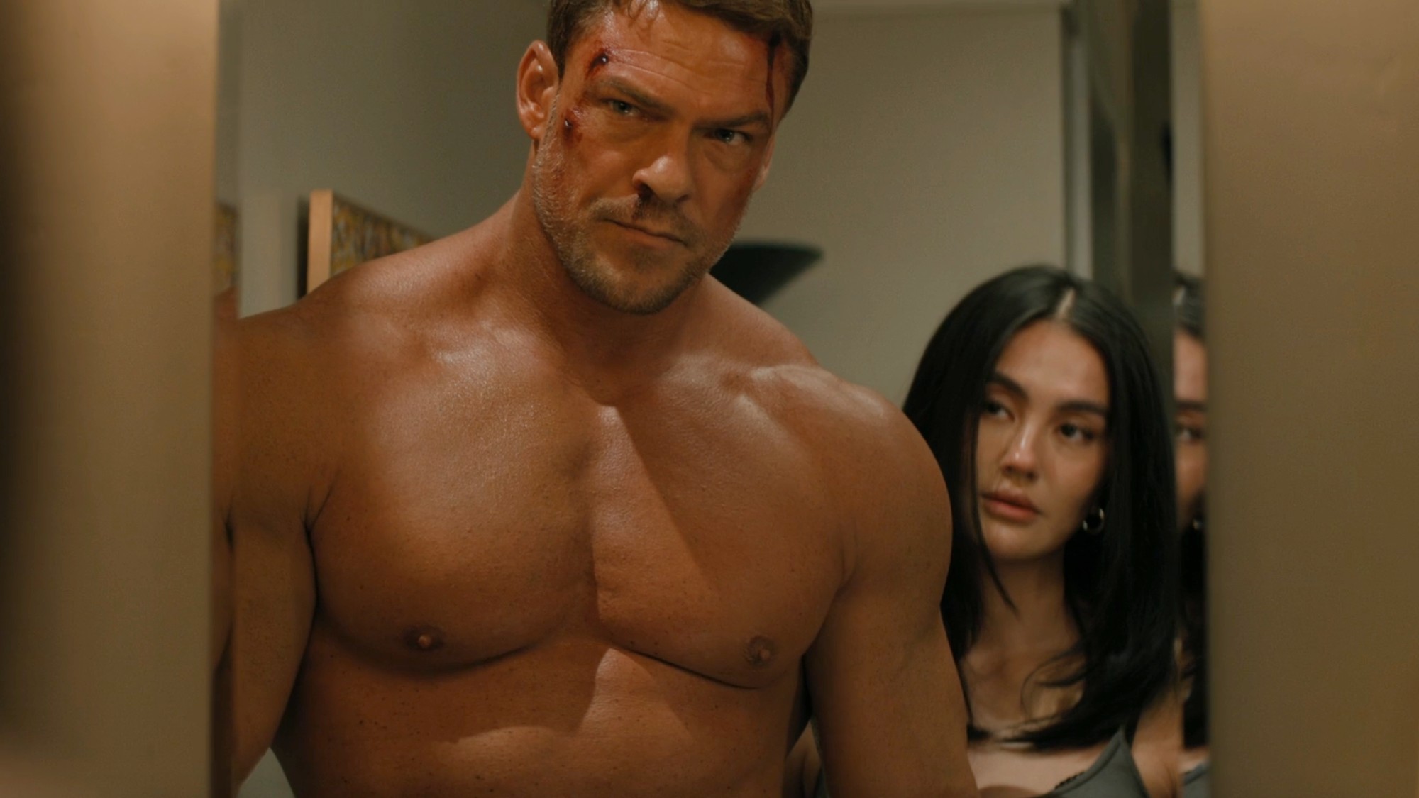 Alan Ritchson shirtless in Reacher while Agnez Mo stands behind him. 