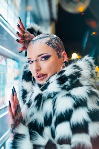 Person with tattooed hands and arms wearing a black and white fur coat, leaning against a window