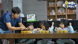 a man (left) tries to feed three babies sitting at a long desk, in the korean show 'return of superman'