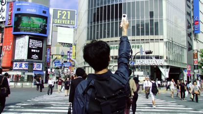 从后面看，一名男子站在东京繁忙的十字路口，手里拿着一个白色的设备指向天空