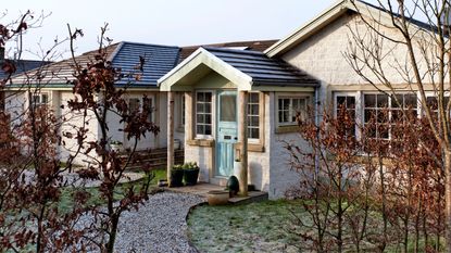 Winter front garden outside exterior of 1980s single story house with gravel path, blue front door and brown-leaved shrubs in the foreground