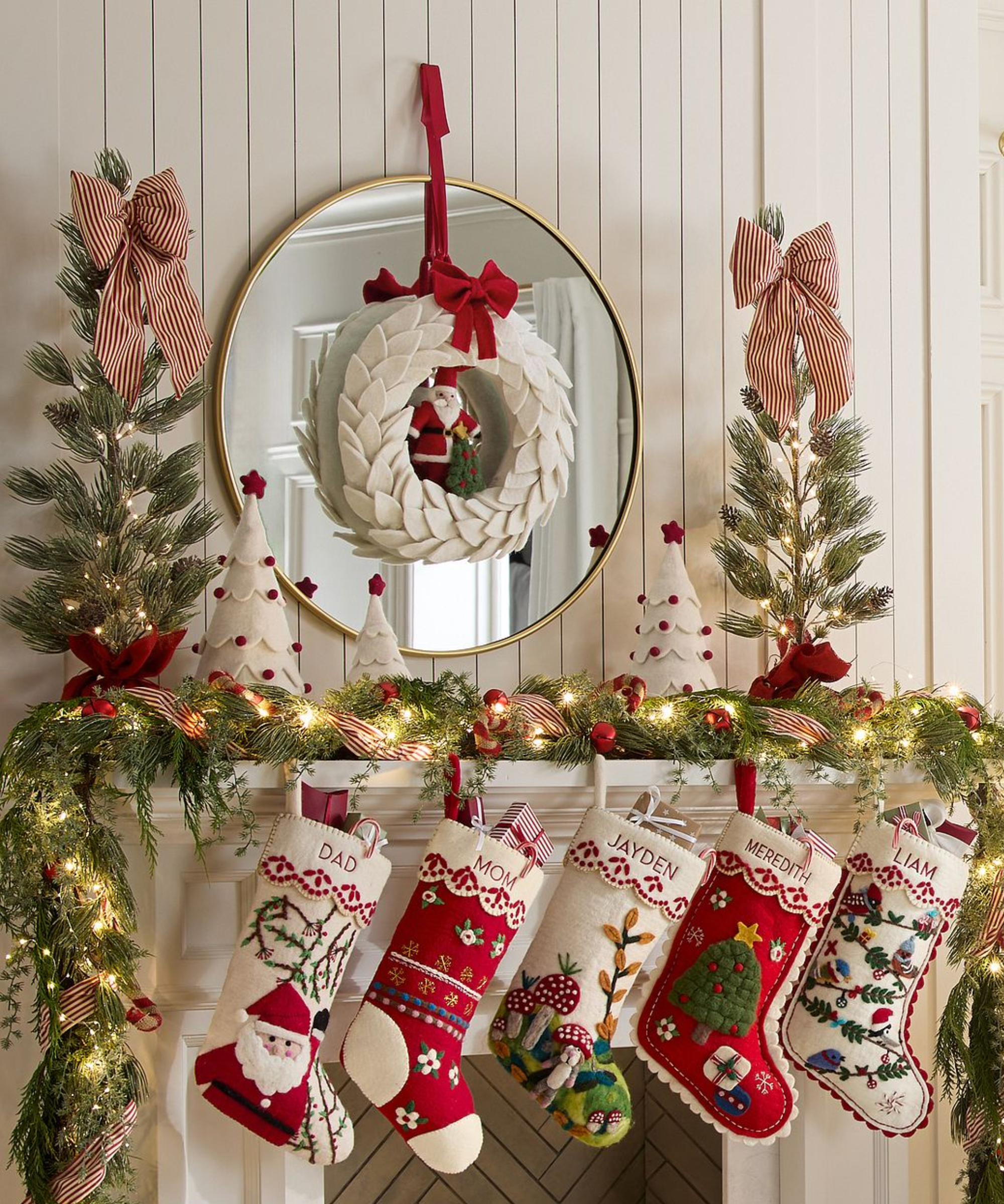 a Christmas display with a mantel and hanging stockings, garland, decorative trees on top of the mantel, and a wreath hanging from a mirror