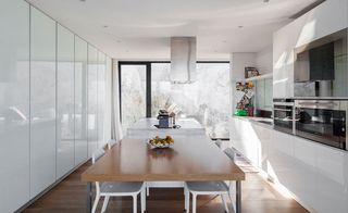 Dining and Kitchen space featuring white walls, white ceiling, brown flooring and floor to ceiling clear glass panel windows. A brown dining table (laid with a bowl of fruits) with white chairs with a white island on the end. White kitchen cabinets and white ceiling lamp