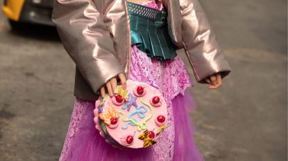 A street style image of a woman wearing a frilly pink skirt and holding a clutch bag shaped like a cake, reminiscent of the best gourmand perfumes