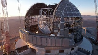 a large domed structure is under construction, with the metal framework of its body shaping to an open ceiling observatory. Cranes stand on either side.