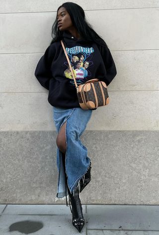 A woman wearing a long denim skirt styled with black knee-high boots, a black sweatshirt, and a vintage bag.