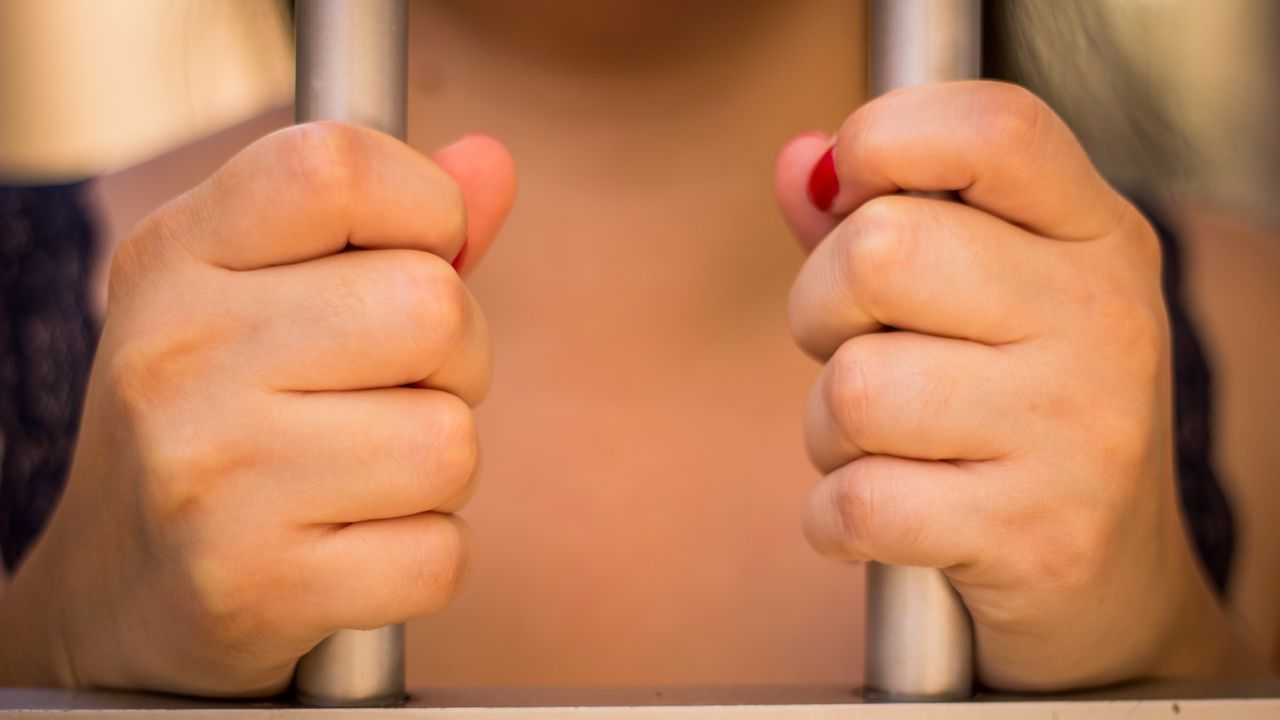 A woman&#039;s hands grip the bars of a jail cell.