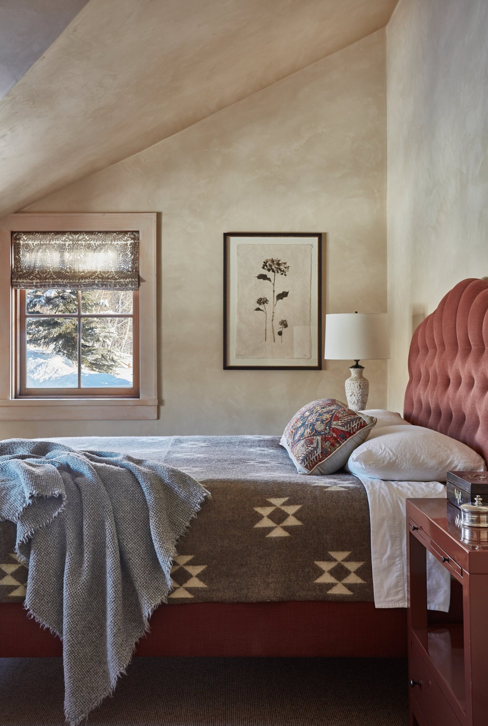 modern bedroom with sloped ceiling, limewash walls, red upholstered bedframe with artwork on wall, vintage-style quilt, timber side tables and throw draped over the end of the bed