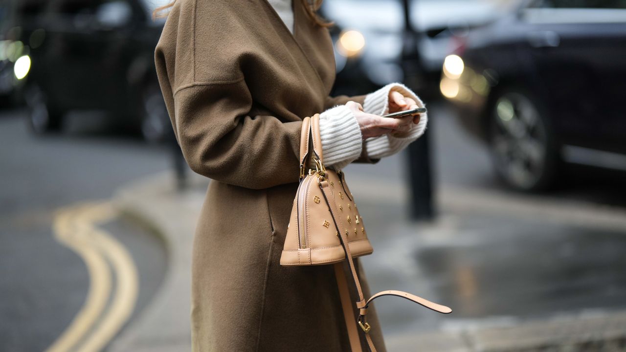  A guest wears a white turtleneck pullover , a brown long coat, a beige and brown tie and dye print pattern midi dress, a brown leather with embroidered gold metallic monogram pattern Alma handbag from Louis Vuitton, outside David Koma, during London Fashion Week February 2023 on February 18, 2023 in London, England. 