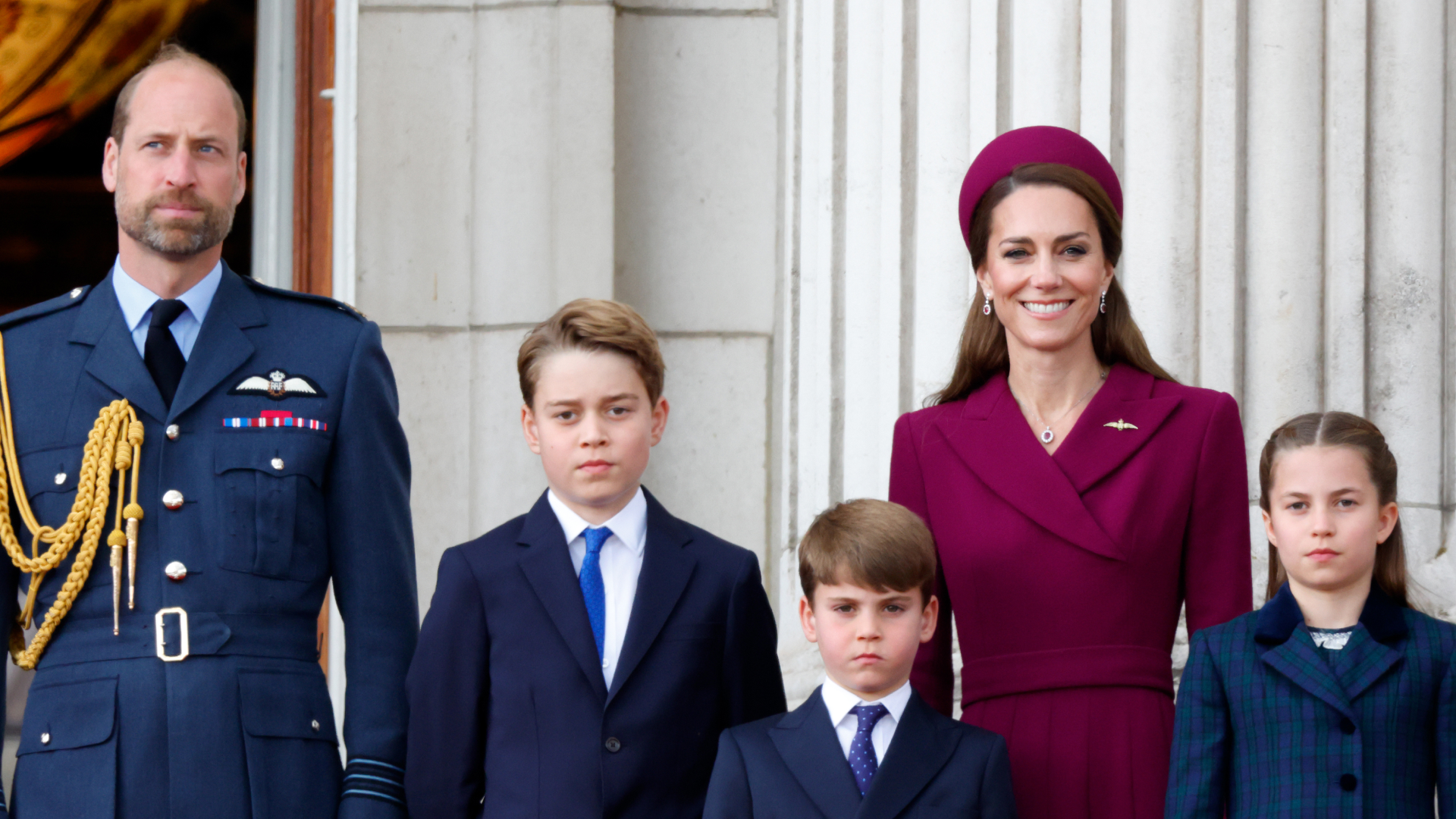 Prince William wears military uniform and Kate Middleton wears a dark red coat dress and matching hat as they stand with children George, Charlotte, and Louis on the Buckingham Palace balcony