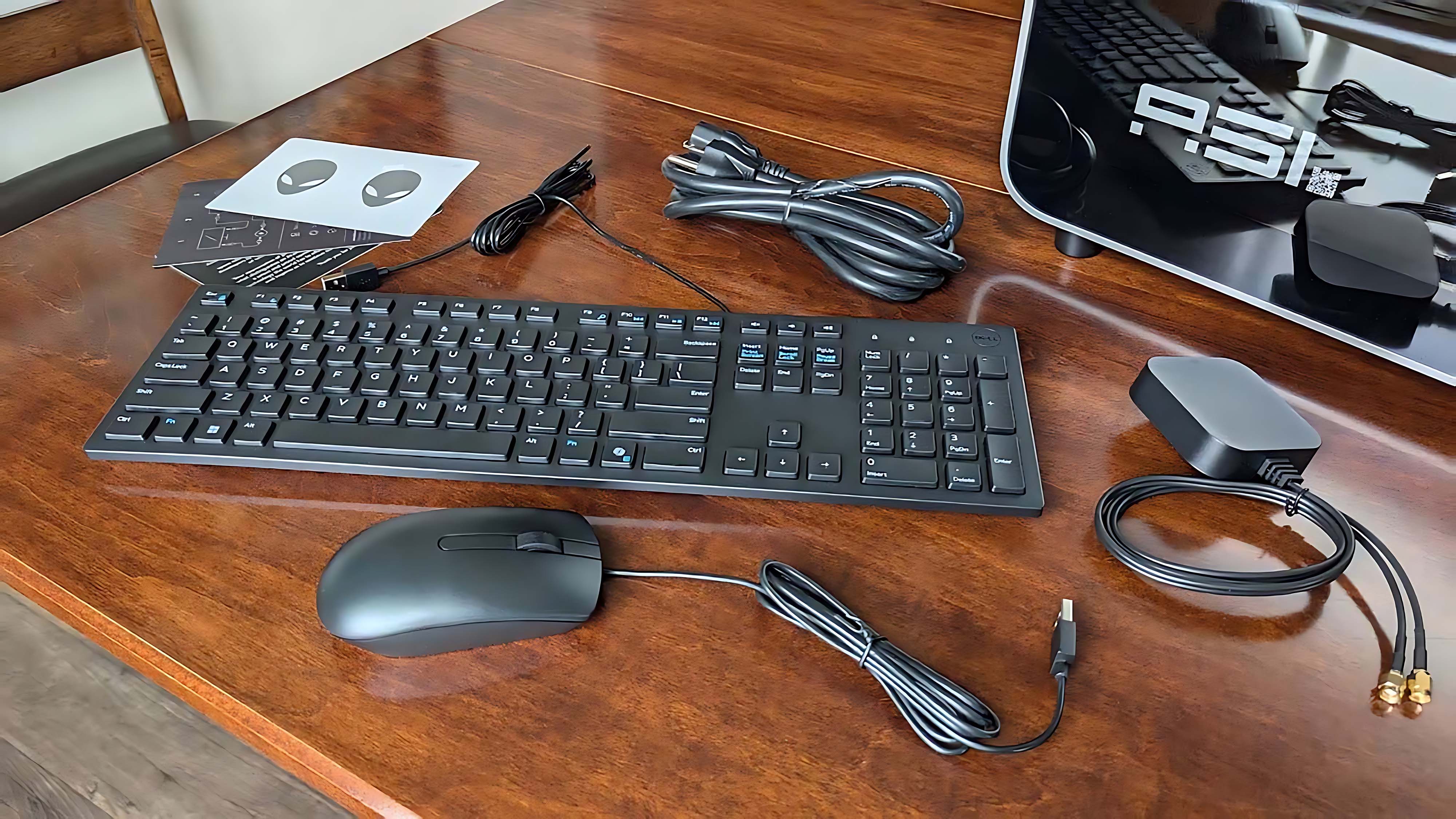 The included mouse, keyboard, Wi-fi antenna, cable, and manuals on a wooden table next to the Alienware Area-51 Gaming Desktop (2025).