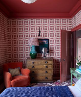 bedroom with burgundy/red ceiling, cornice and woodwork, red patterned wallpaper, dresser, lamp, red armchair, rug, green side table, bright blue throw on bed