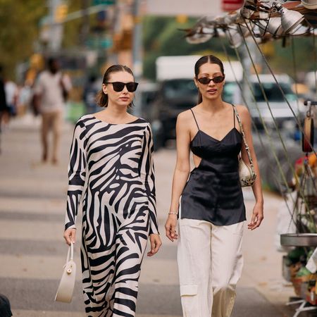 Two women are walking in the streets of NYC. One is wearing a zebra jumpsuit, and the other is wearing a black satin top with white pants. 