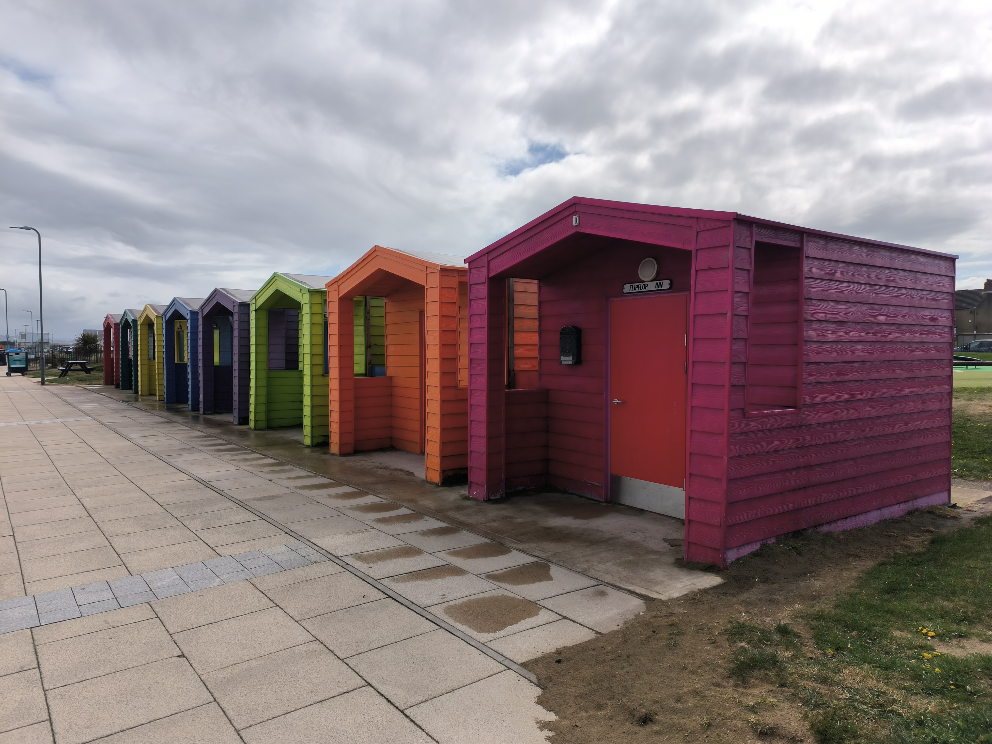 Row of brightly painted beach huts in various colours—pink, orange, green, purple, blue, and yellow—lined up along a paved promenade with grass and cloudy skies overhead