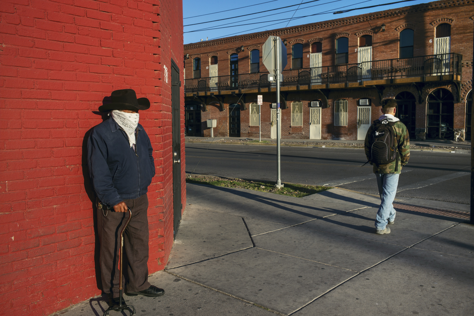 An older man in a black cowboy hat leans against a bright red brick wall, while a young man in a camouflage jacket and backpack walks away down a sunny pavement towards a row of brick buildings.