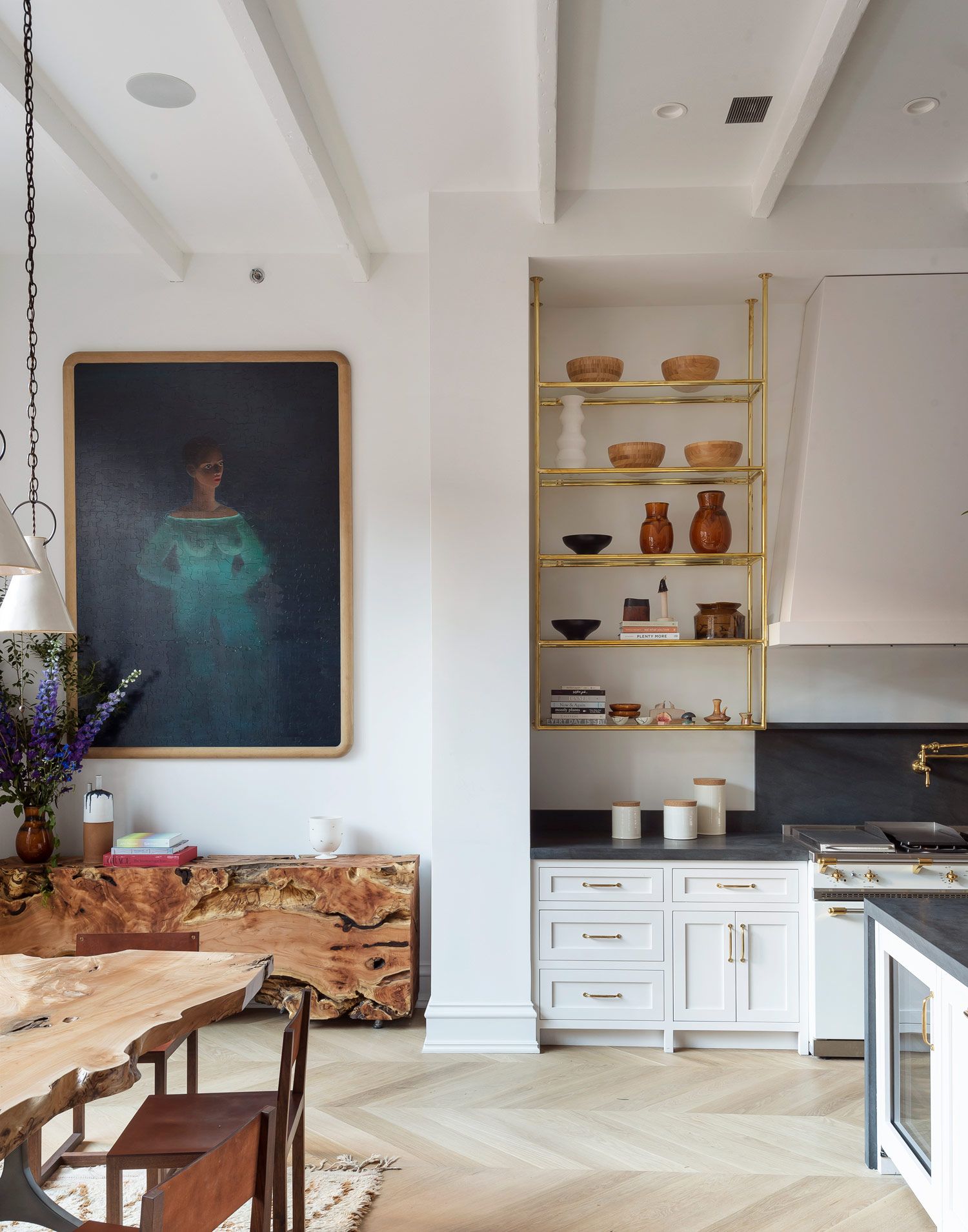 A modern white kitchen and dining space with white walls, wall-hung gold and glass open shelving, a wood dining table, and a wood console under a large, dark painting of a lady wearing a blue dress 