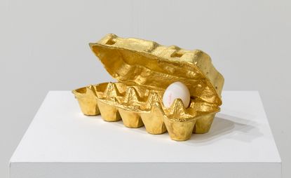 A ten egg box painted in gold, half open with one white egg inside. 