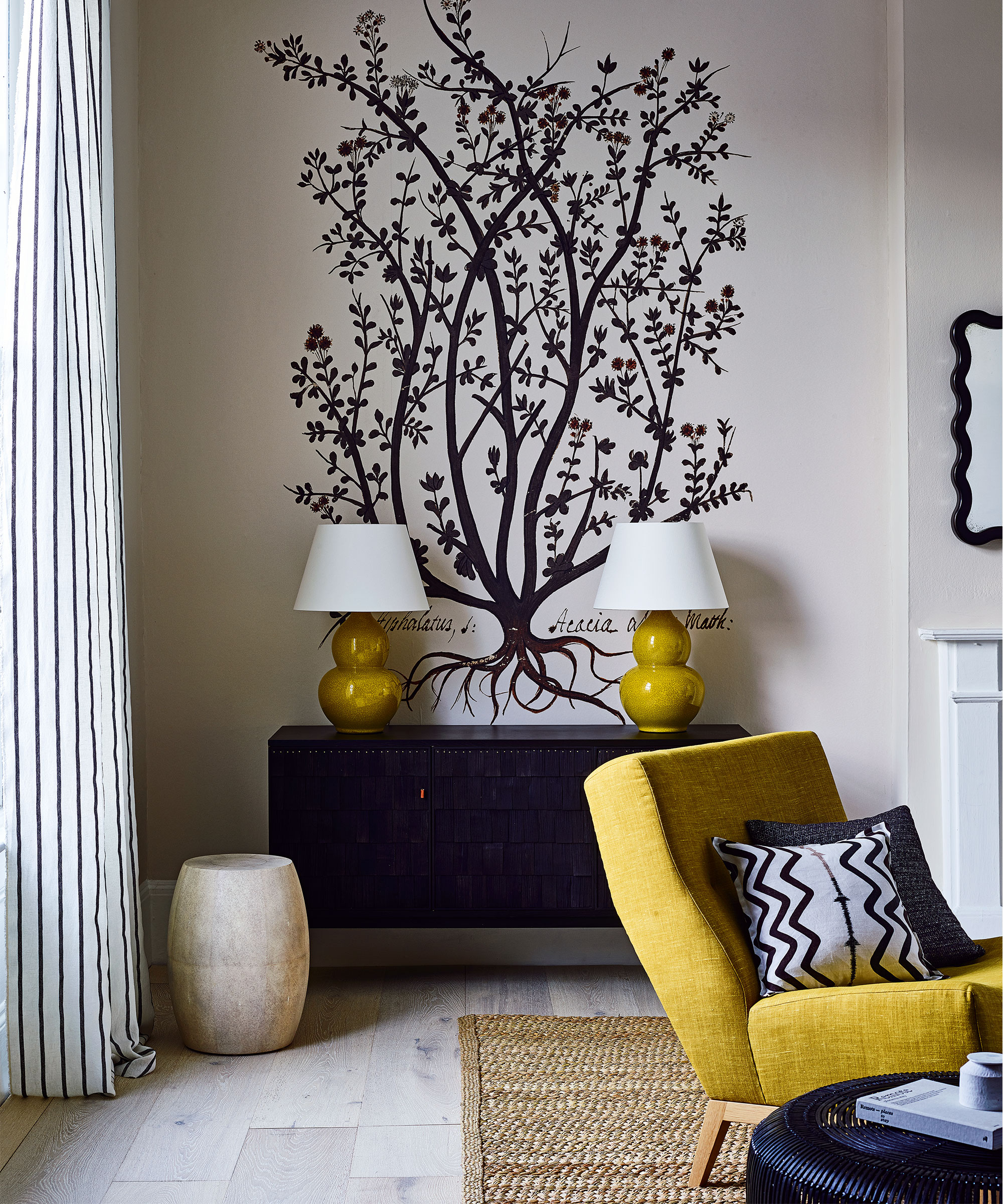 A living room with a yellow armchair and table lamps in front of a white wall with a black tree motif