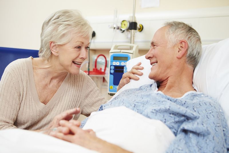 An older couple talks while the man lays in a hospital bed