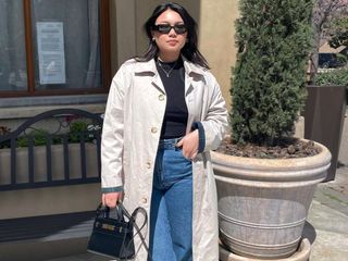 female fashion influencer Marina Torres poses outside a building wearing black oval Prada sunglasses, a black mock-neck top, light beige trench coat, high-waisted jeans, and a mini black YSL bag