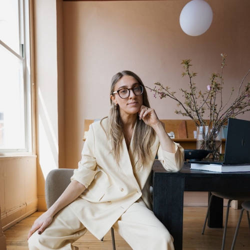 Image of a woman in a pale yellow suit with long hair and glasses. She is sitting at a desk in a beige room.