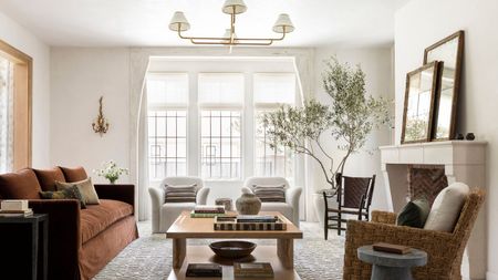 A light and airy living room with white walls, large windows, a rust-colored sofa and a large wood coffee table with books and decor. A textured rug and two cream armchairs. 