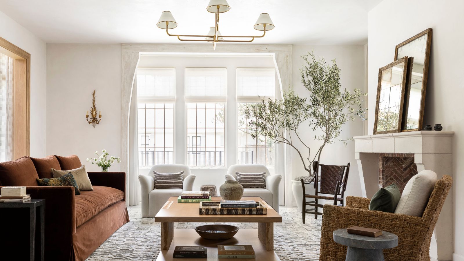 A light and airy living room with white walls, large windows, a rust-colored sofa and a large wood coffee table with books and decor. A textured rug and two cream armchairs. 