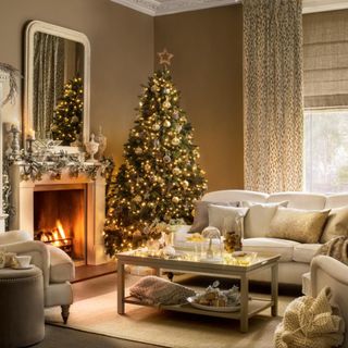 Decorated Christmas tree in a country style brown living room, white sofa, cushions, fireplace, lit fire, decorated mantelpiece, large mirror, coffee table.