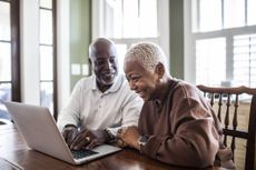 An older, smiling couple makes plans over a laptop in an elegant kitchen or dining room.