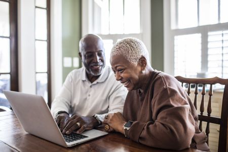 An older, smiling couple makes plans over a laptop in an elegant kitchen or dining room.